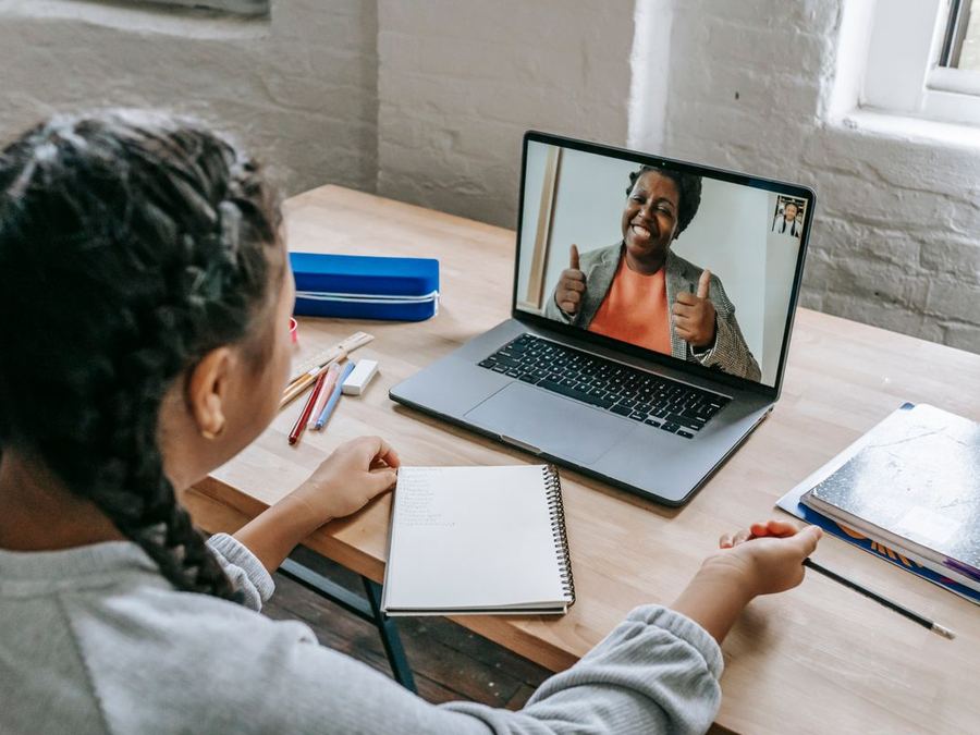 A student at a desk taking notes while a tutor explains a topic on a laptop video call