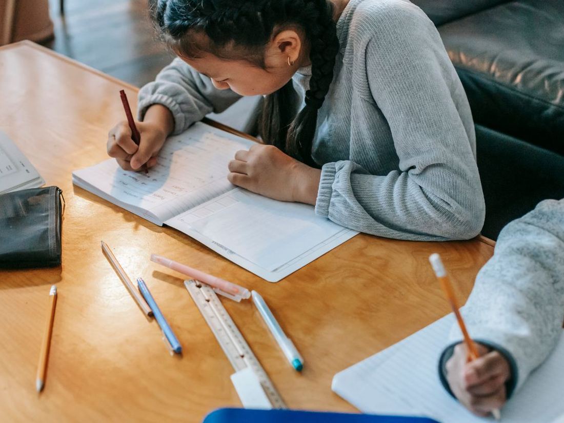 Two students writing in exercise books at a wooden table, pencils and rulers beside them